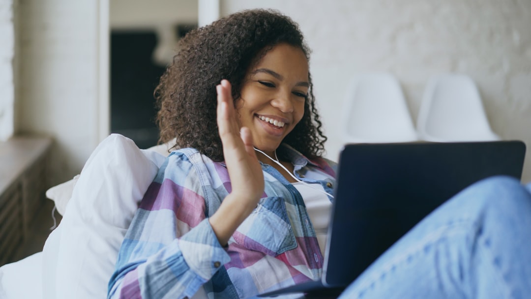 Cheerful mixed race girl having video chat with friends using laptop camera while lying on bed at home